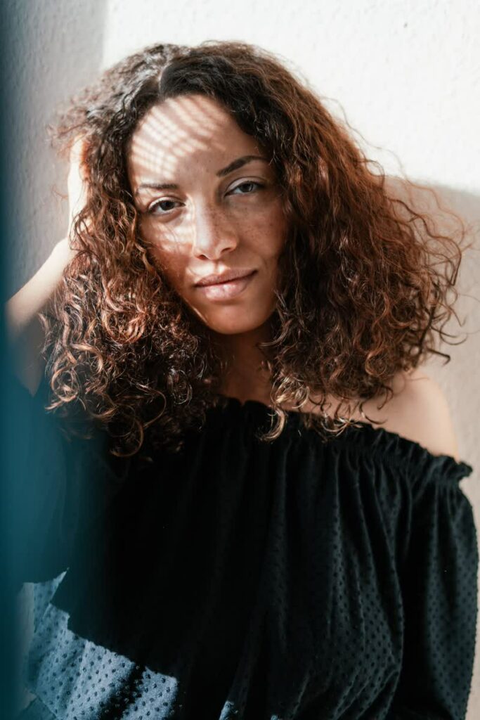 eine Frau mit braunen lockigen Haaren an einem Fenster, der Sonnenschein scheint auf ihr Gesicht