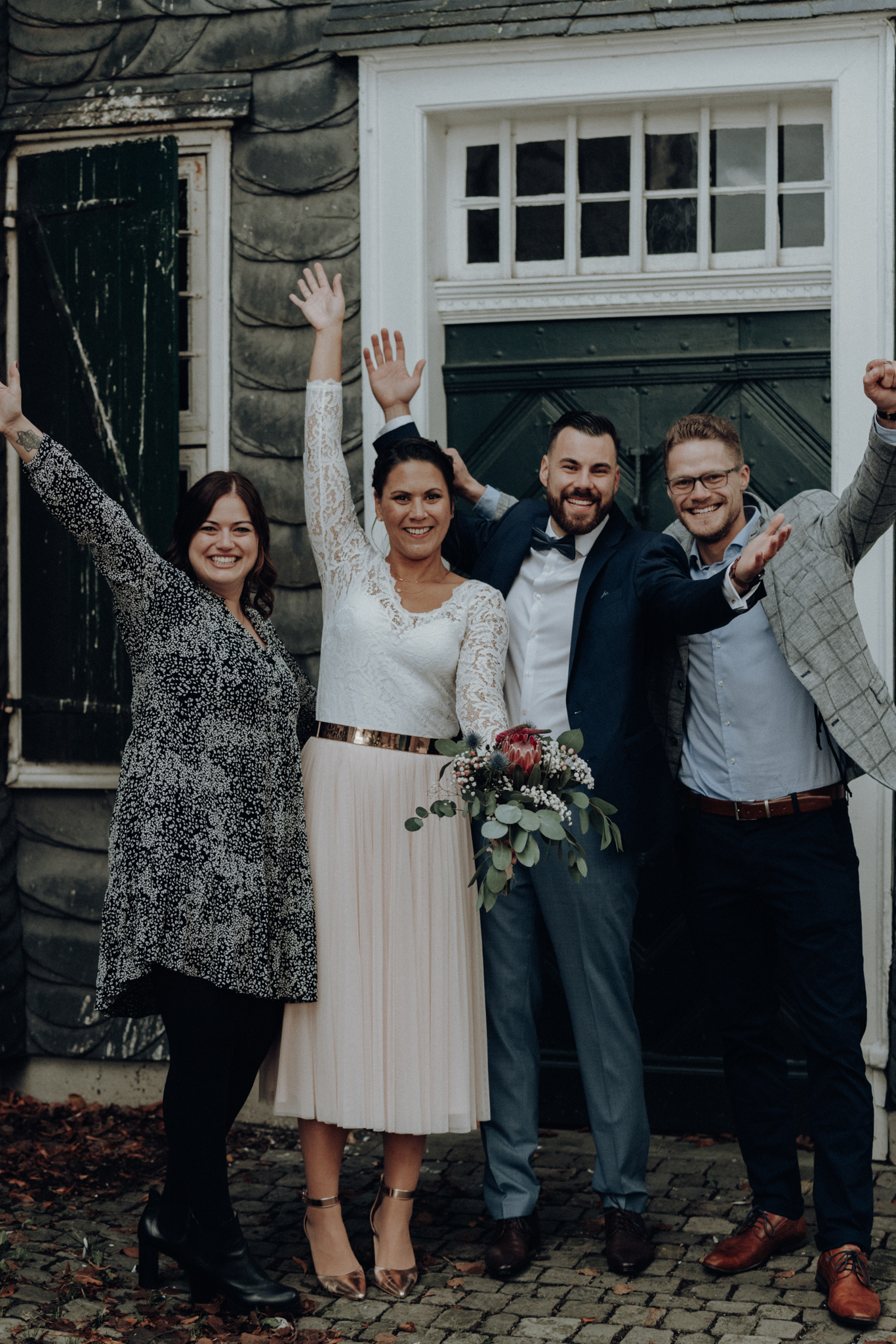 Hochzeitspaar steht mit Freunden vorm Standesamt und streckt die Hände in die Luft