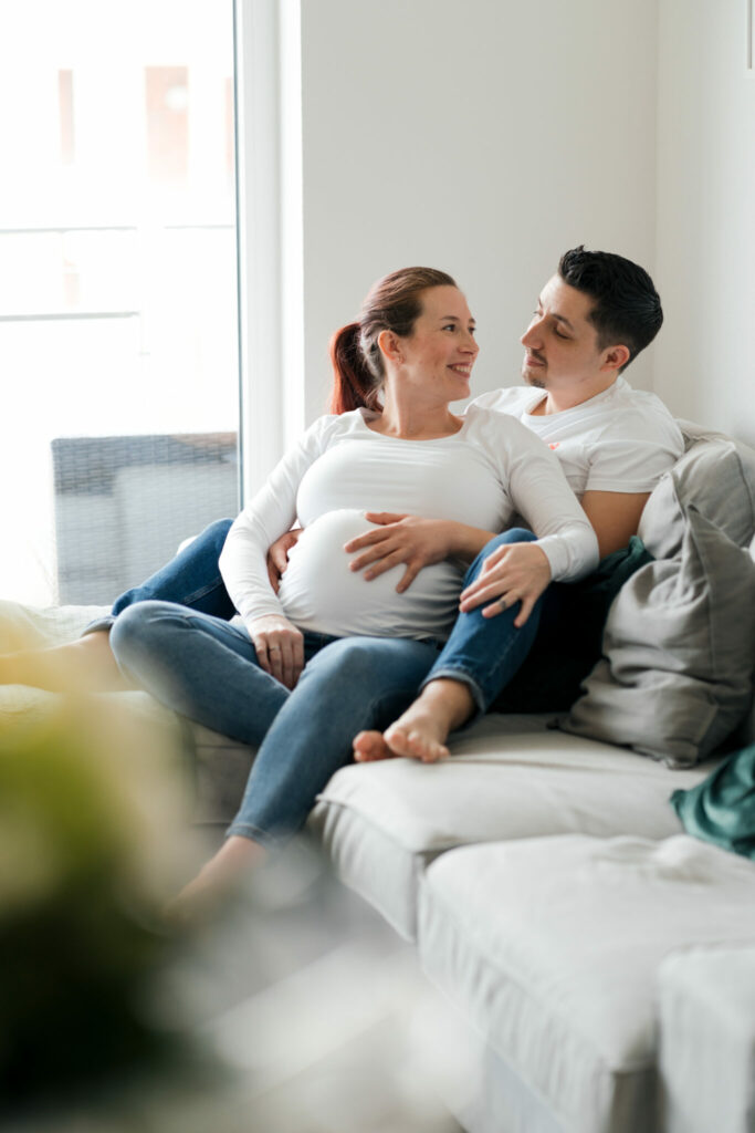 Schwangere Frau sitzt lächelnd im Schoß von ihrem Freund auf dem Sofa