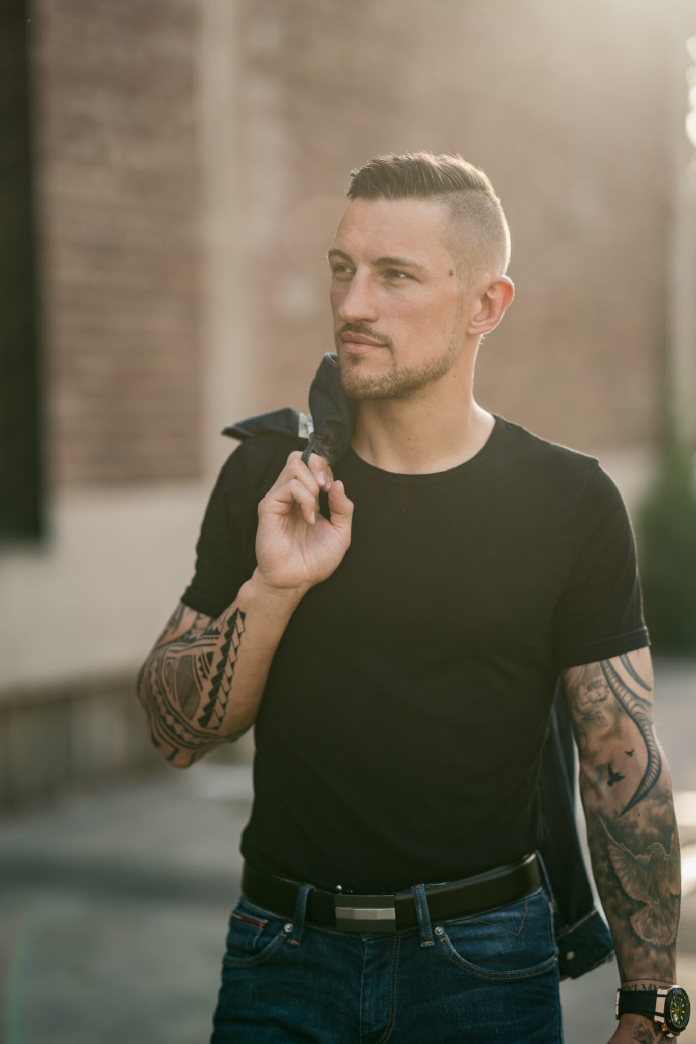 Junger Mann mit Bart und Tattoos steht bei Sedcard-Shooting vor Hauswand und schaut zur Seite