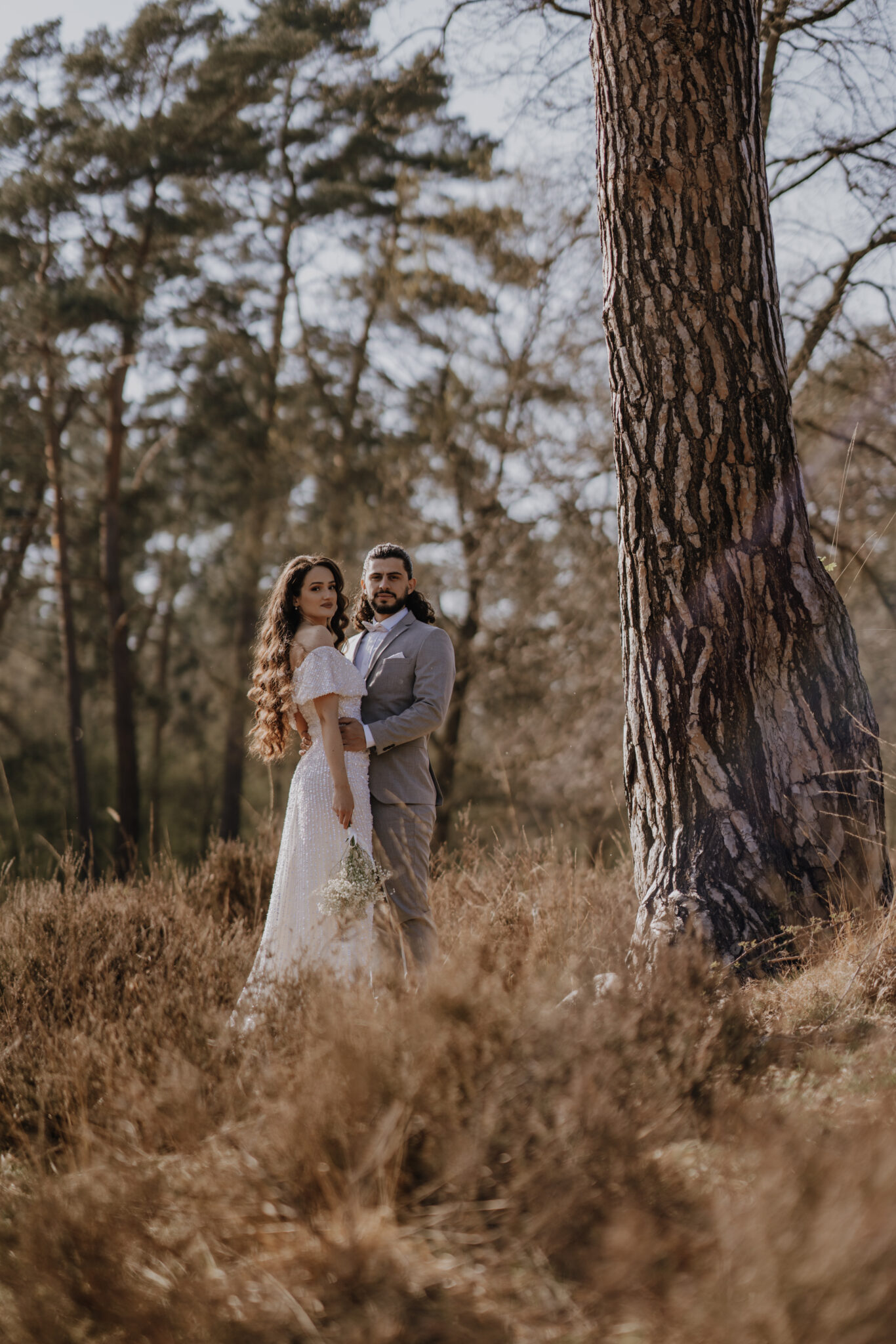 Das Brautpaar steht neben einem grToßen Kieferbaum, beide gucken in die Kamera und der Bräutigam hält die Braut an ihrer Taille