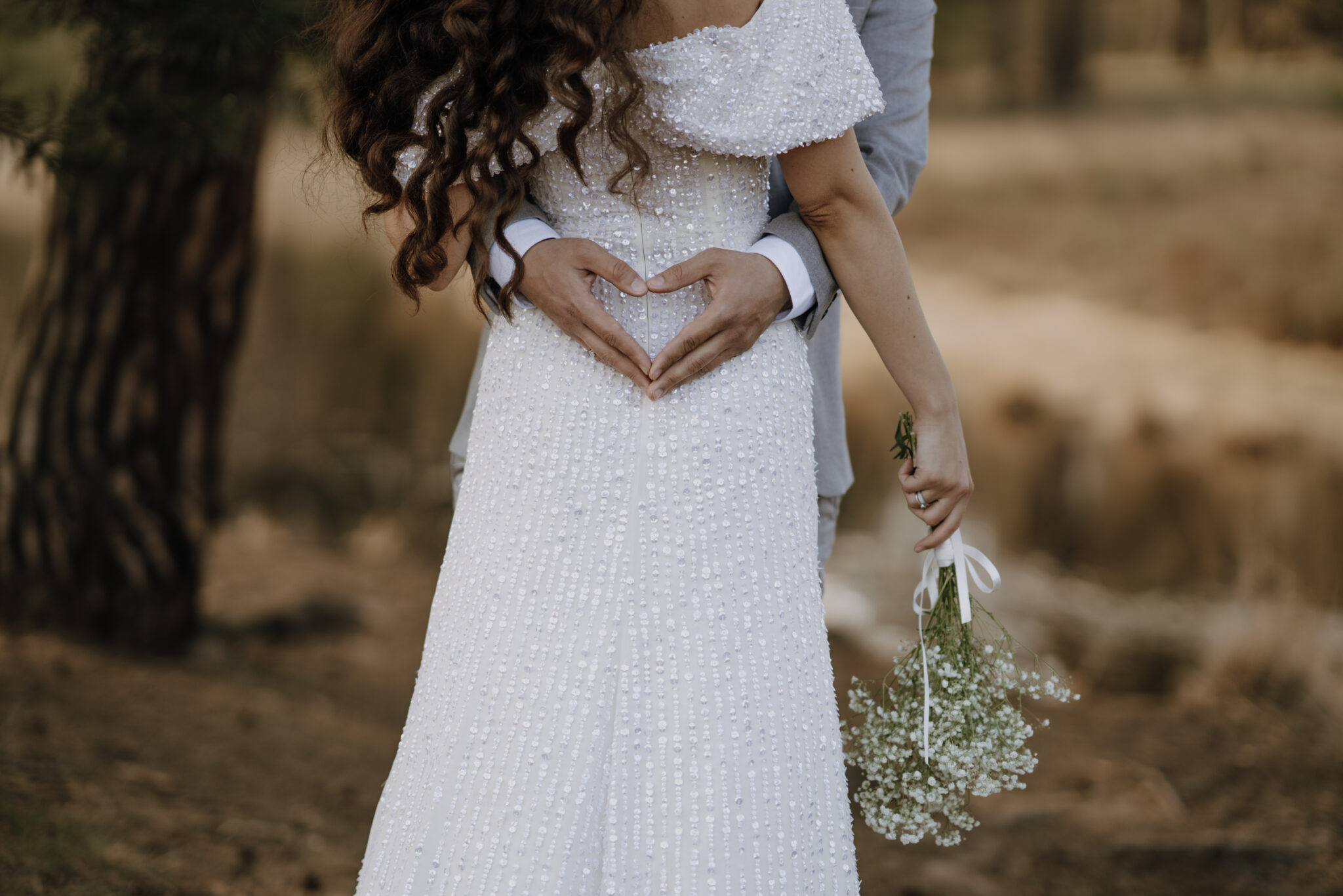 Die Braut steht mit dem Rücken zur Kamera. Der Bräutigam umarmt die Braut und formt die Hände zu einem Herz 