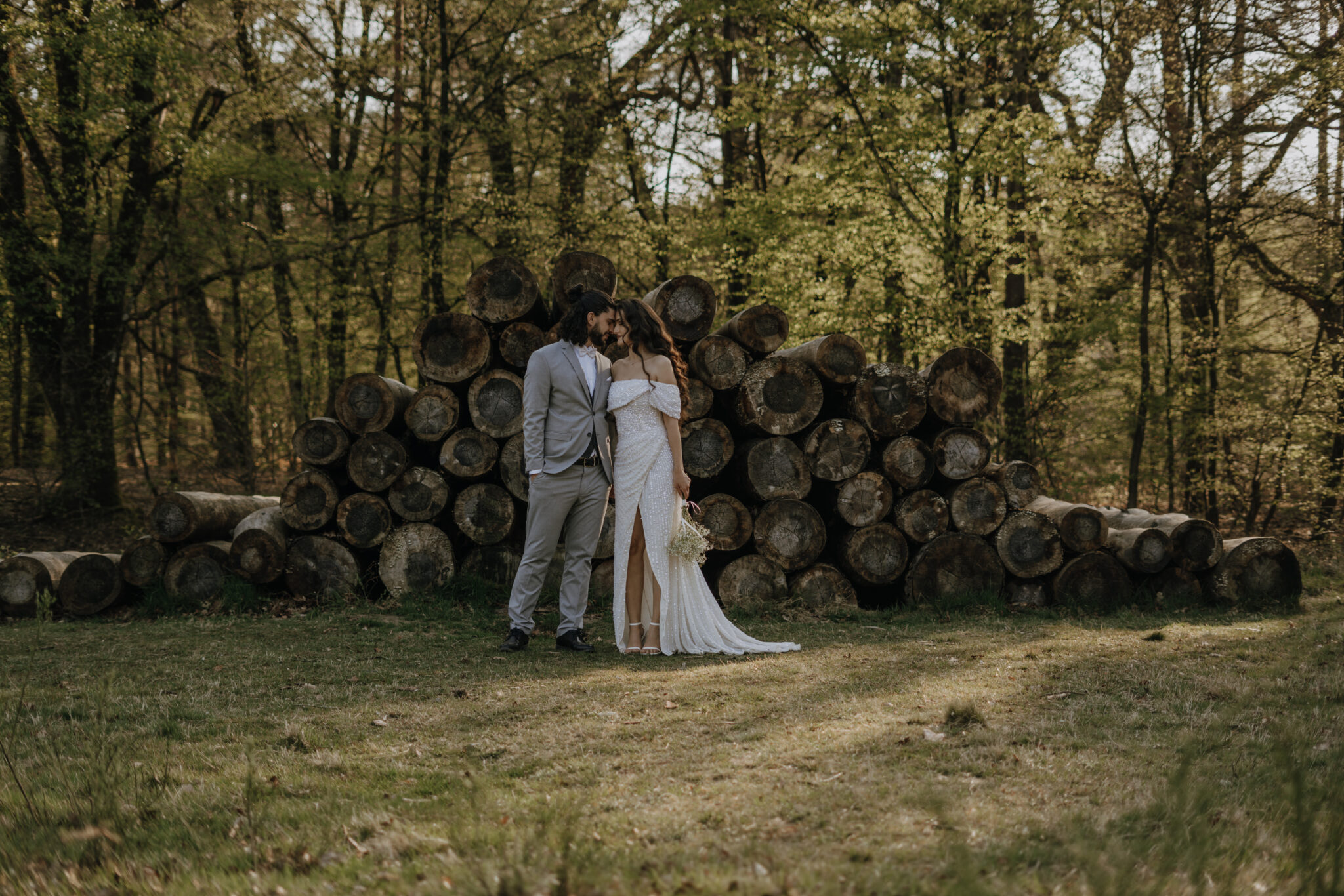 Bei diesem Hochzeitsfoto, steht das Brautpaar nebeneinander vor gestapelten, abgeholzten Baumstämmen und schmiegen die Köpfe aneinander währenddessen die Augen der beiden geschossen sind