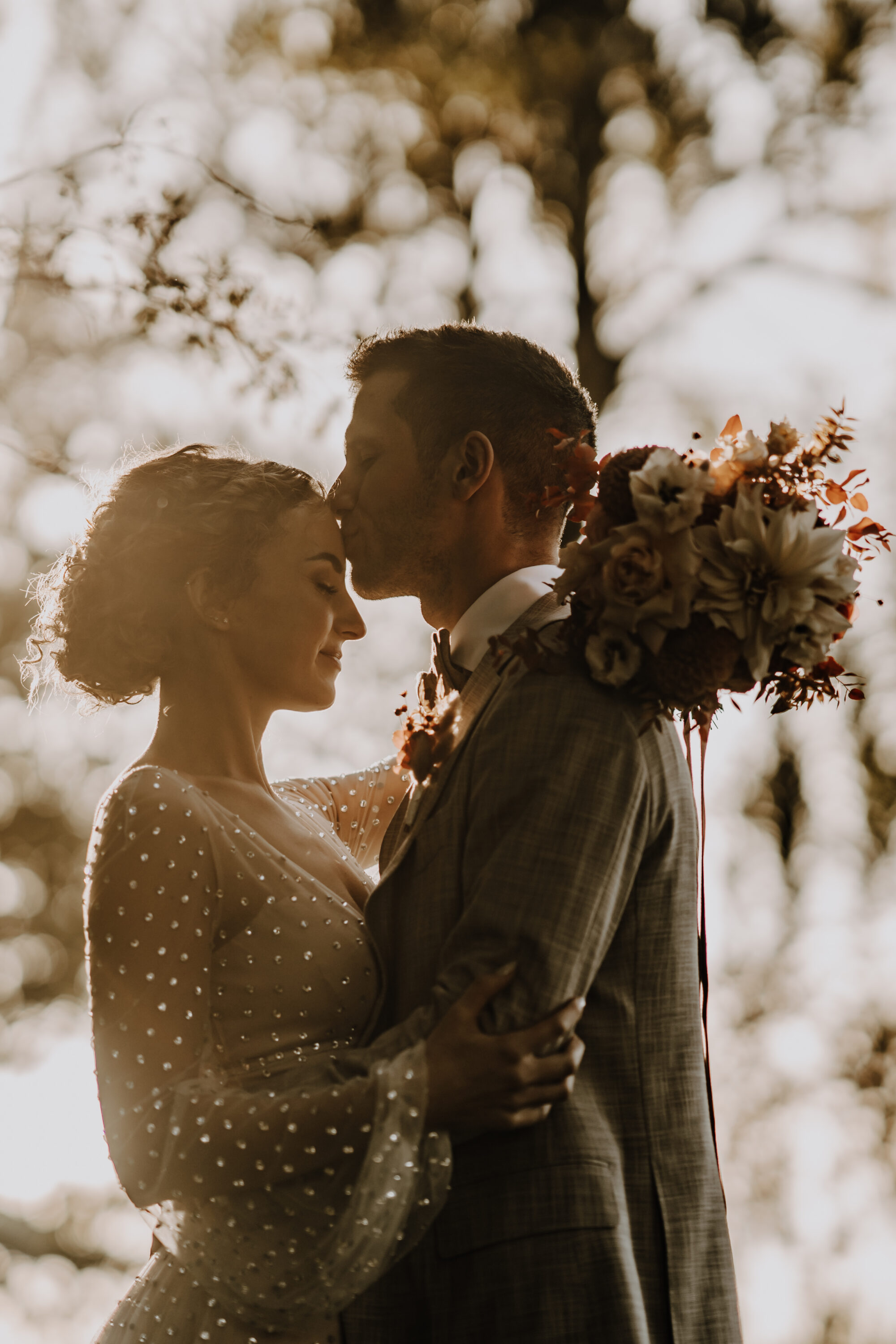Hochzeitspaar, sich im Arm haltend, gegen Sonne, warmes Licht, Bräutigam küsst Braut auf Stirn
