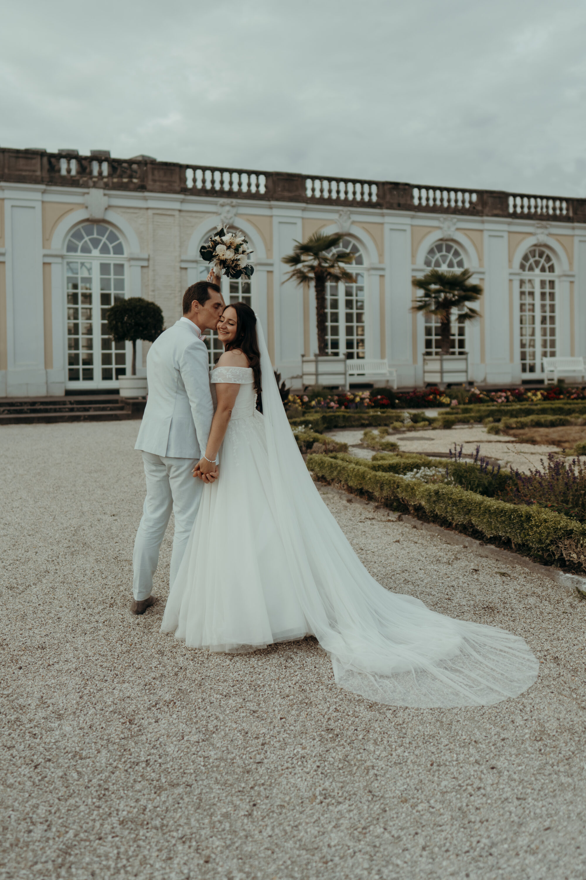 Hochzeitspaar in Schlossgarten, Bräutigam küsst Braut auf Wange, Braut hält Strauß hoch