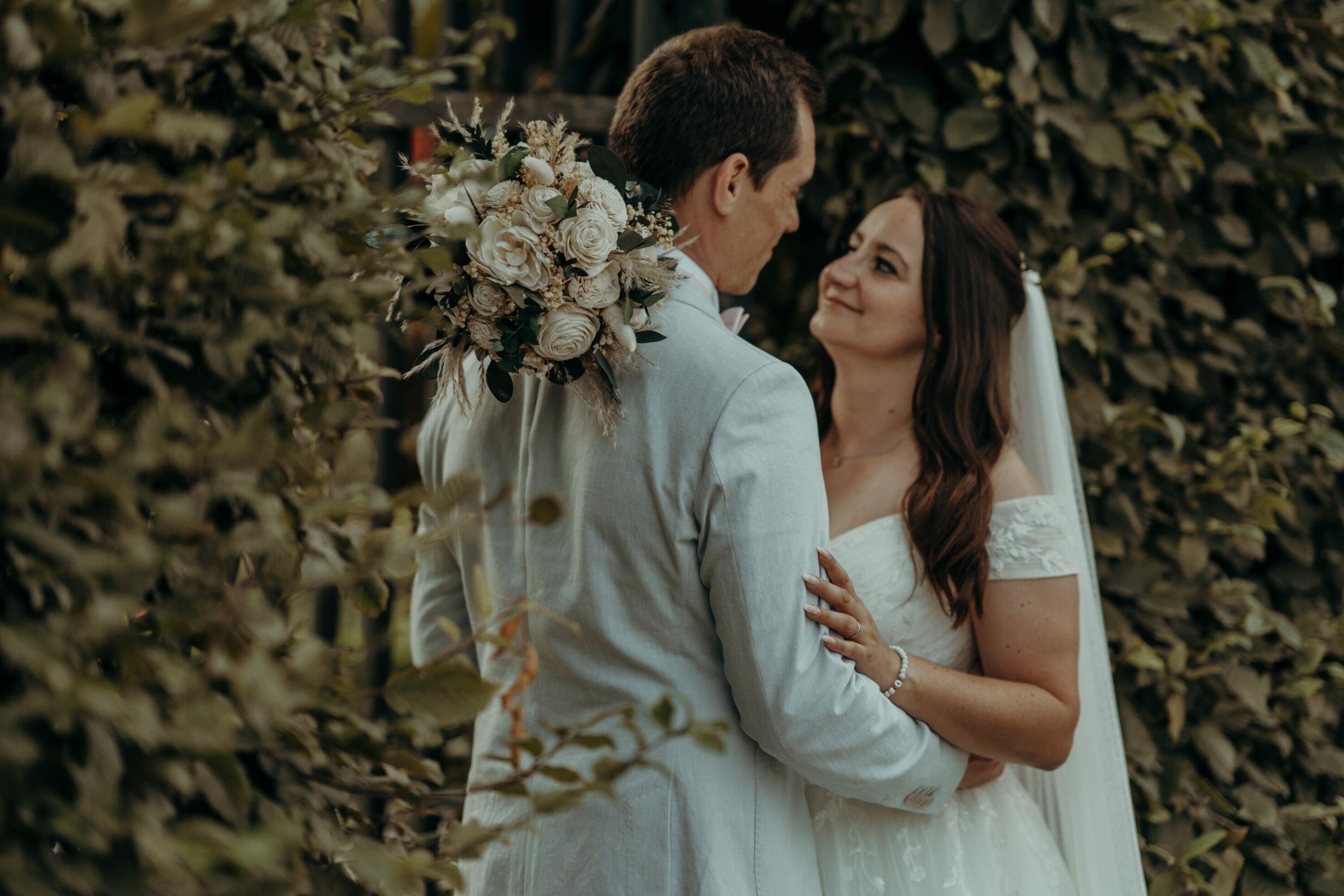 Brautpaar schaut sich in die Augen, gerahmt von Hecken, Brautstrauß fokussiert auf Schulter des Bräutigams