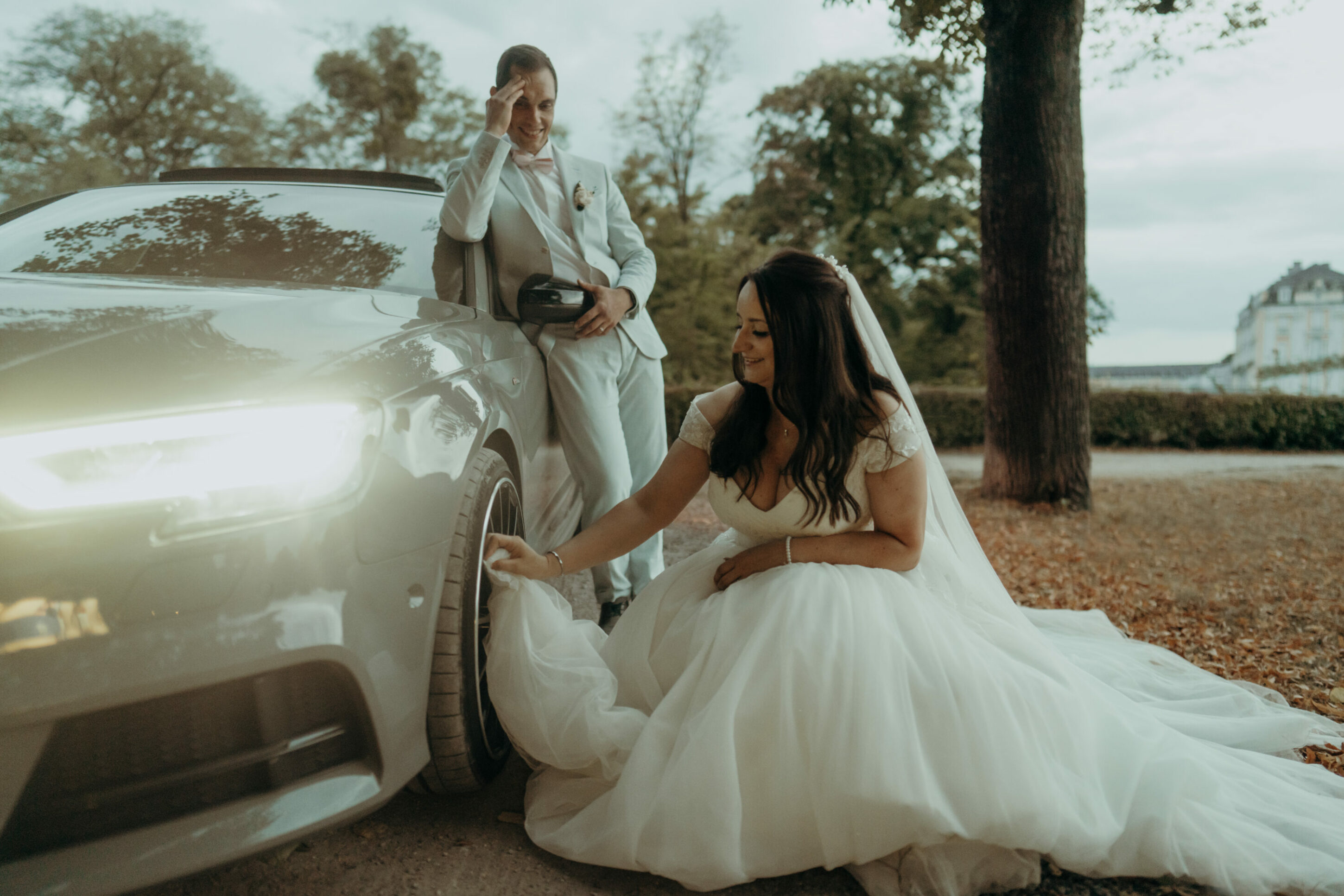 Braut putzt Felge des Autos mit Kleid, Bräutigam lacht und fasst sich an die Stirn