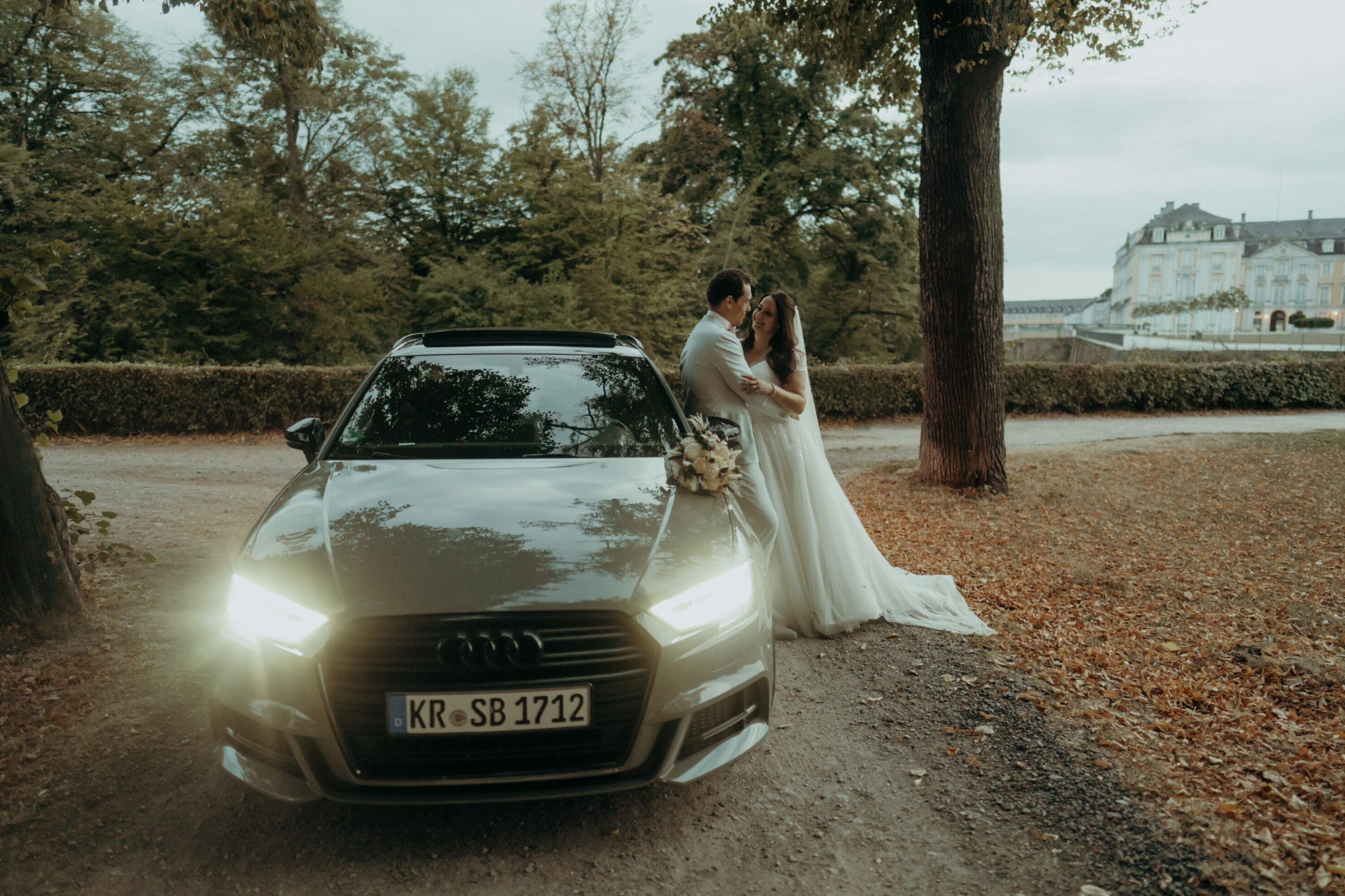 Hochzeitspaar lehnt an Audi, sich im Arm haltend, Schloss im Hintergrund, Allee