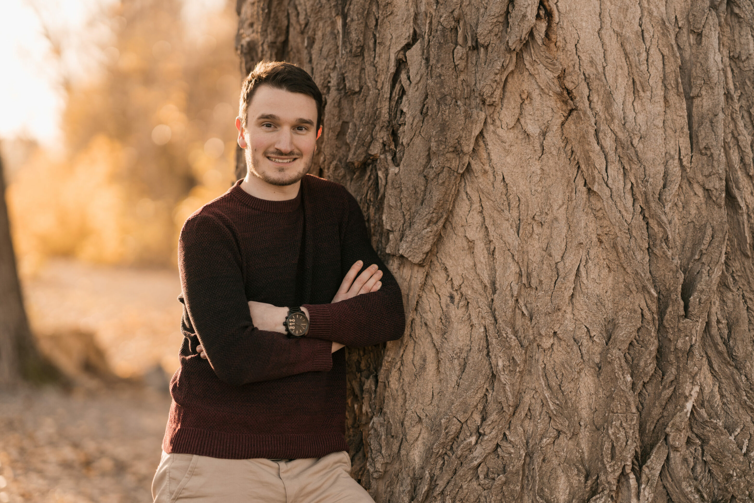 Junger Mann steht im Herbst an Baum gelehnt mit verschränkten Armen und lächelt 