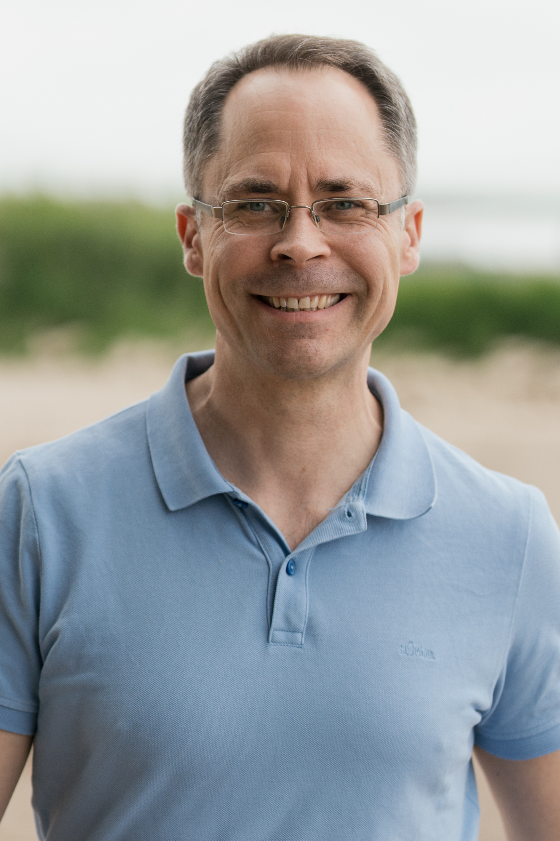 Mann mit Brille steht gerade, im Hintergrund Strand und lächelt in die Kamera