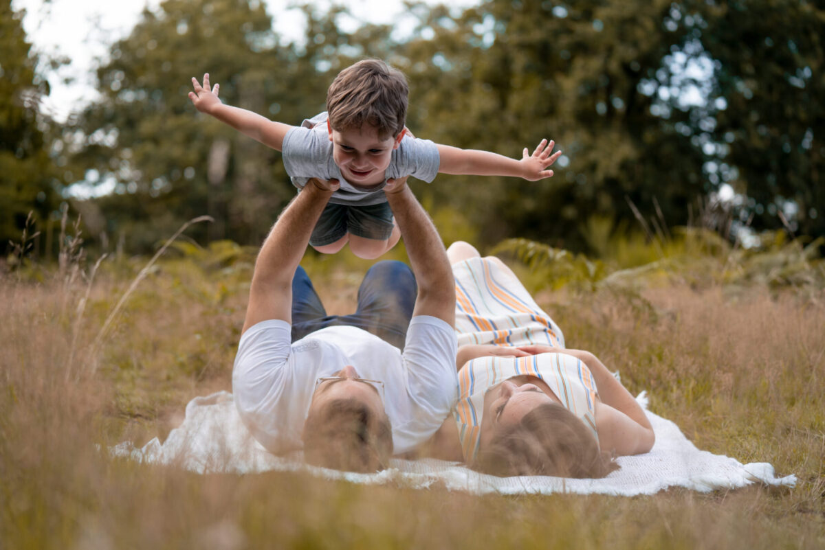 73R00223-2judith Familienfotoshooting, Eltern, die mit ihrem Sohn den Flieger machen, während sie in der Wahner Heide auf dem Boden liegen.