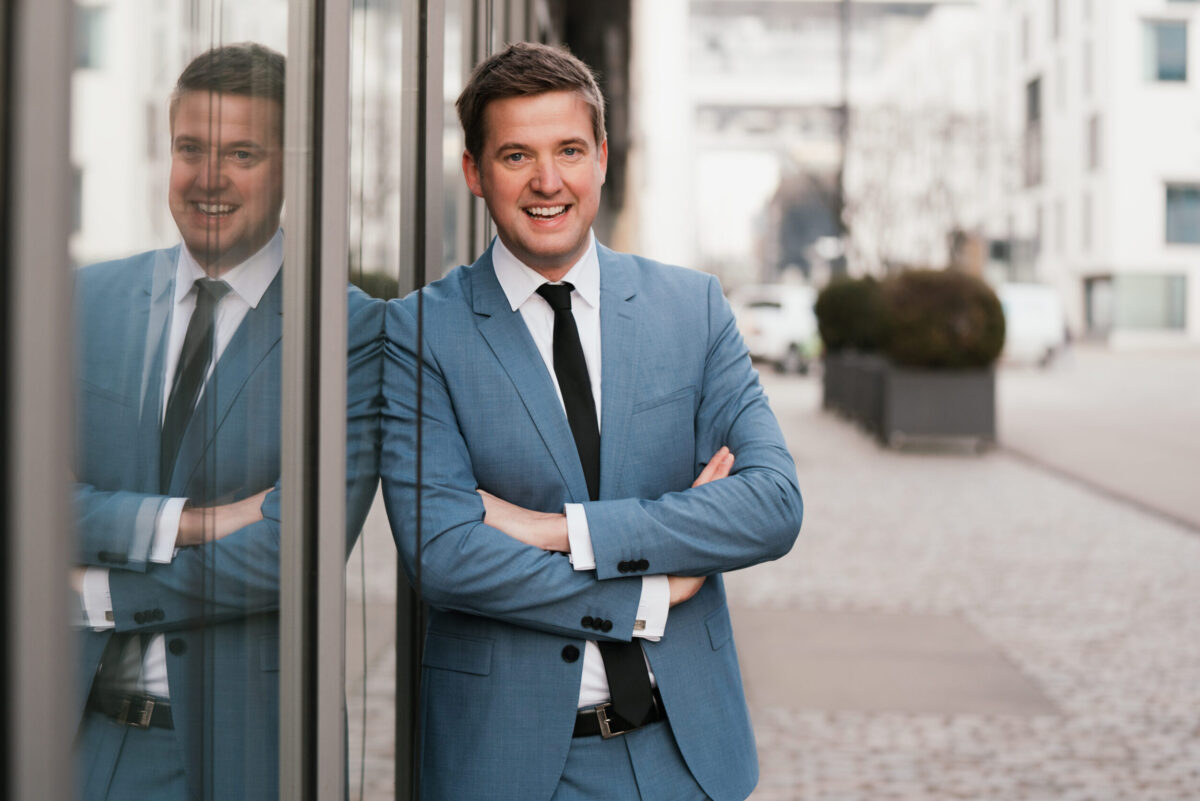 Businessfoto von einem Mann im blauen Anzug, der angelehnt an einer Glaswand steht und mit verschränkten Armen in die Kamera lächelt.