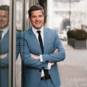 Businessfoto von einem Mann im blauen Anzug, der angelehnt an einer Glaswand steht und mit verschränkten Armen in die Kamera lächelt.
