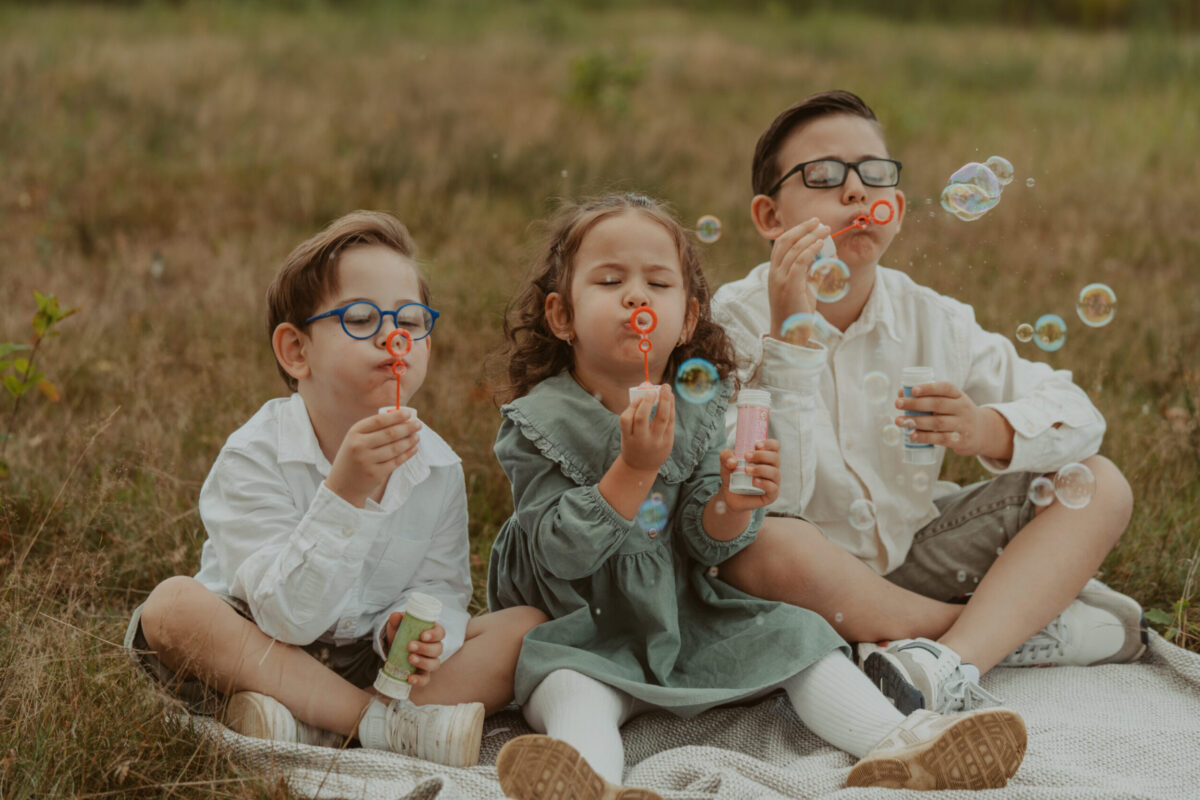 as_familienshooting_102023-_4R20717-2 Kleinkindfotos, bei dem drei Kinder, die im Gras sitzen, Seifenblasen pusten und einfach Kind sind