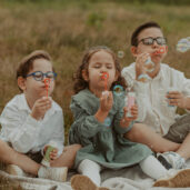 Kleinkindfotos, bei dem drei Kinder, die im Gras sitzen, Seifenblasen pusten und einfach Kind sind