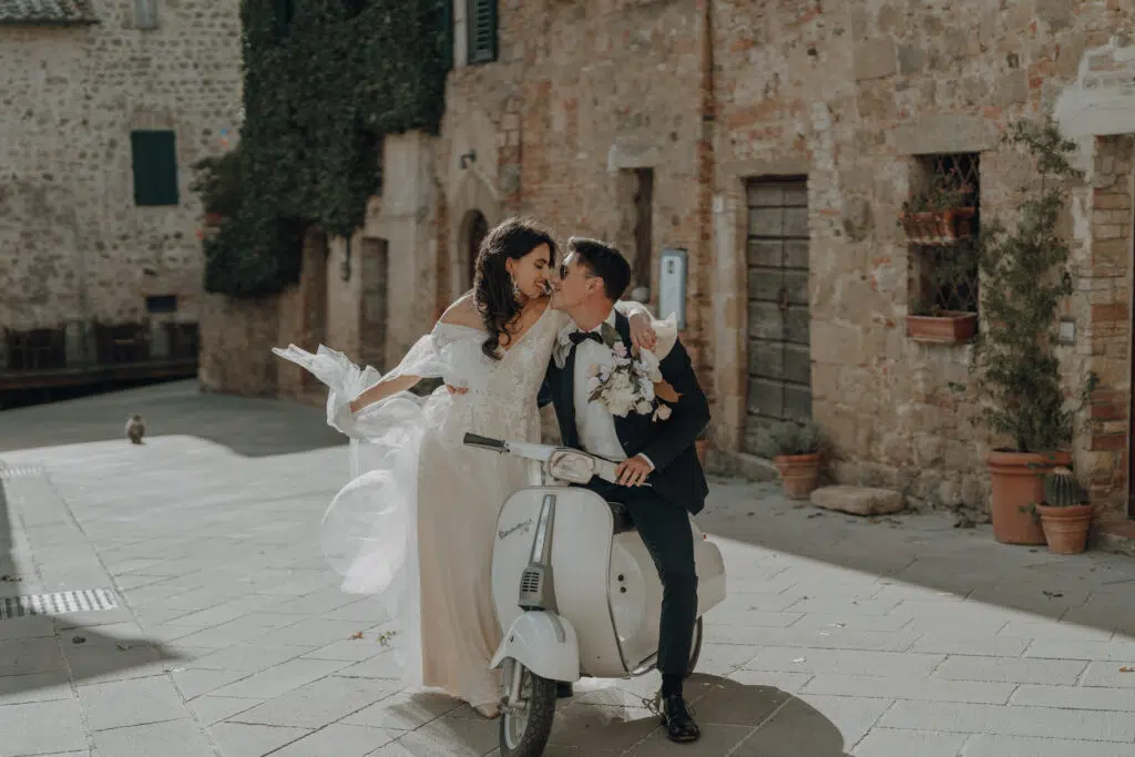 Hochzeitsportfolio, Bräutigam sitzt auf einem Roller die Braut steht daneben und Umarmt ihn von der Seite