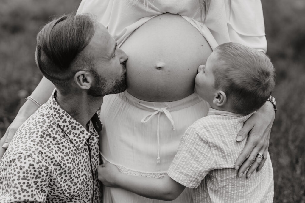 Babybauch Fotografie Portfolio Vater und Kleinkind küssen den schwangeren Bauch der Mutter Schwarz-Weiß-Fotografie