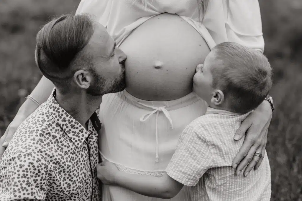 Babybauch Fotografie Portfolio Vater und Kleinkind küssen den schwangeren Bauch der Mutter Schwarz-Weiß-Fotografie