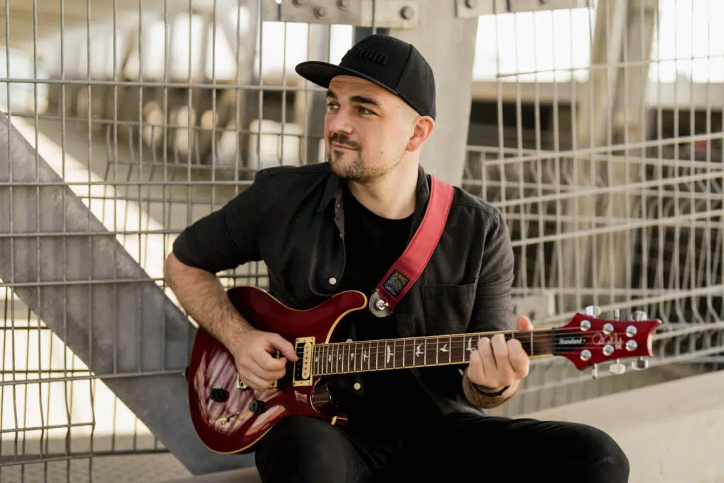 Portraitfoto eines Mannes wie er mit E-Gitarre auf einer Kante sitzt