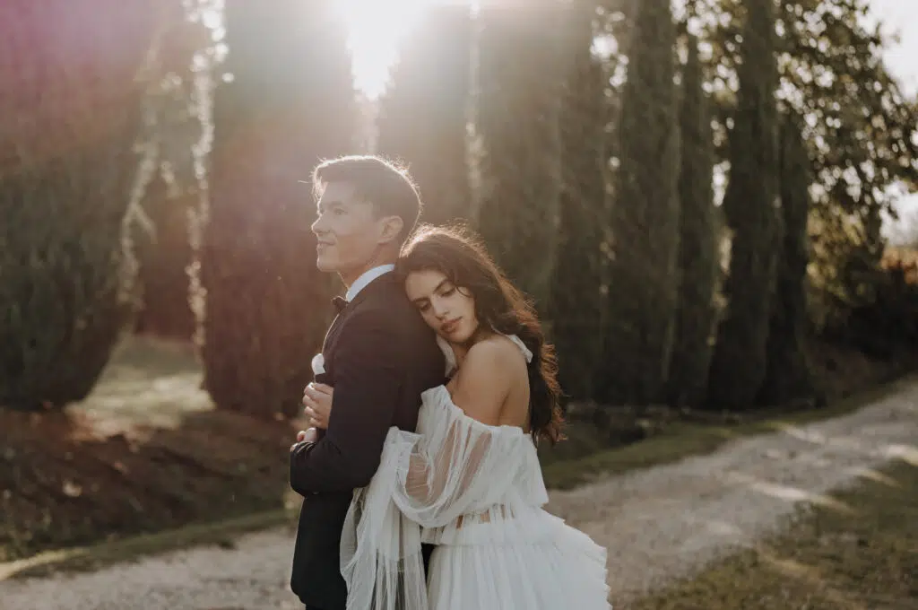 Hochzeitsportfolio, Braut umarmt den Bräutigam von hinten und lehnt ihren Kopf an ihn, Gegenlichtaufnahme im Hintergrund höhe, schmal angeordnete Bäume