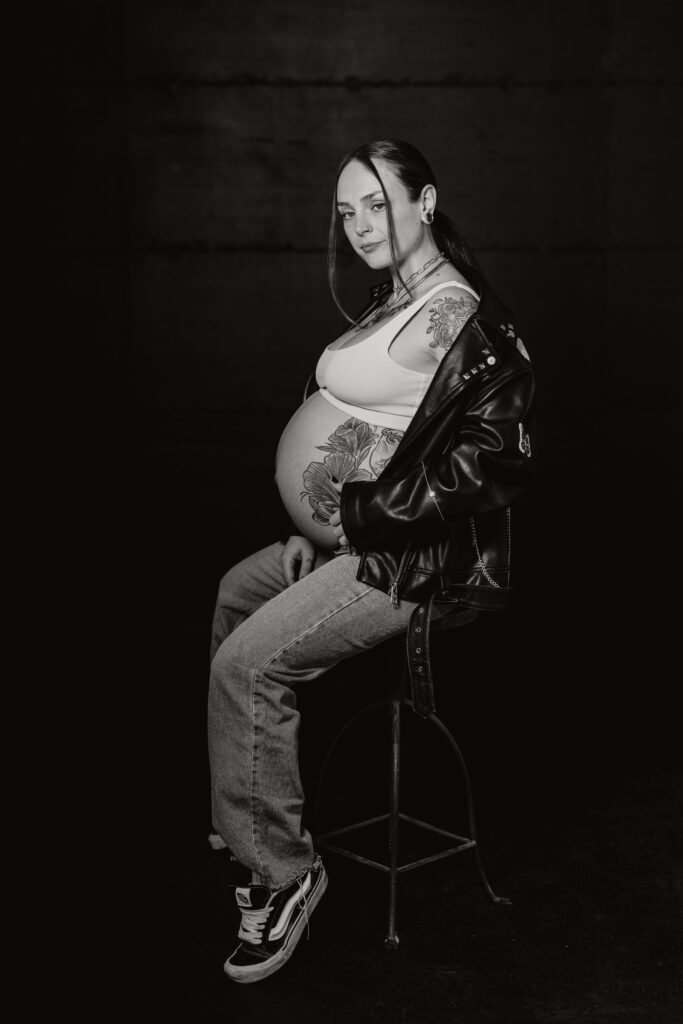 Babybauch Fotografie Portfolio schwangere tätowierte Frau posiert sitzend vor dunkler Wand Schwarz-Weiß-Aufnahme