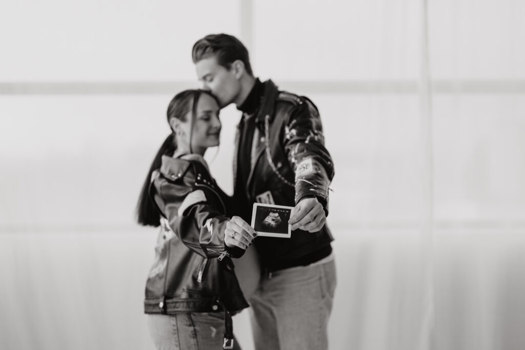 Babybauch Fotografie Portfolio Paar steht zusammen und hält Ultraschallbild in die Kamera Indoor Schwarz-Weiß-Fotografie