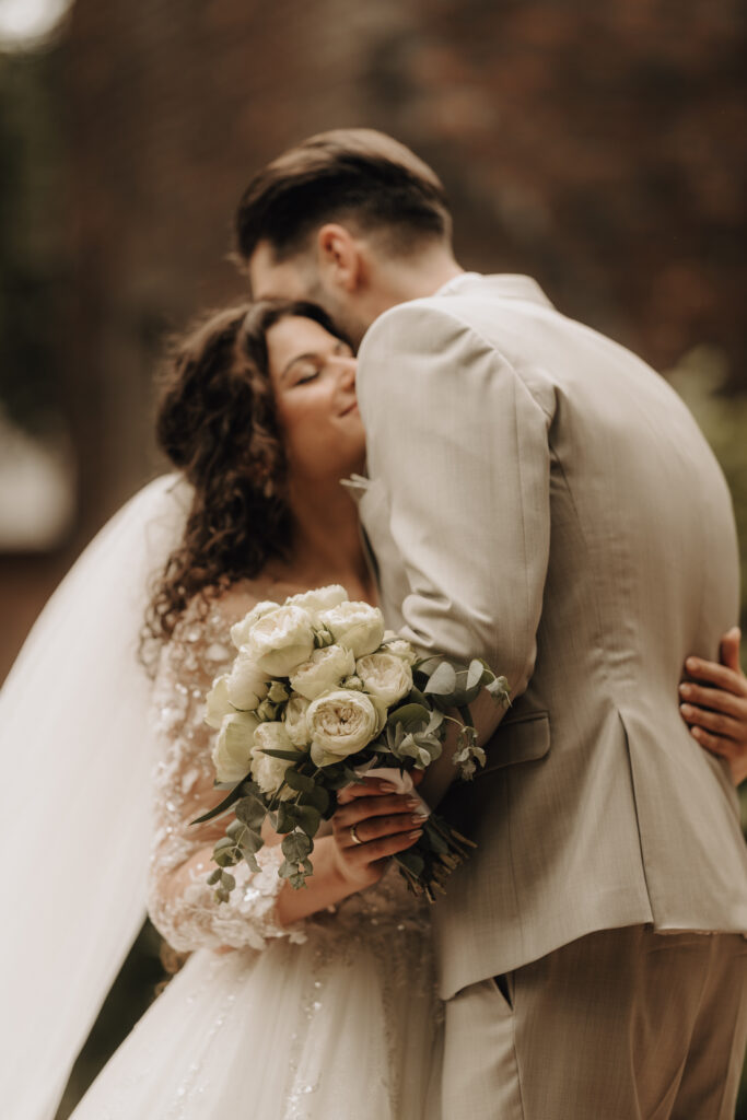 Hochzeitsportfolio, Braut umarmt den Bräutigam von vorne, der weiße Blumenstrauß in ihrer rechten Hand