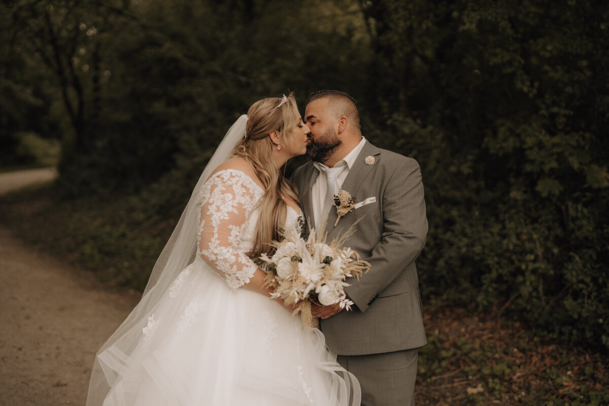 Brautpaar steht vor dem Wald, er küsst sie auf die Stirn, Querformat warm