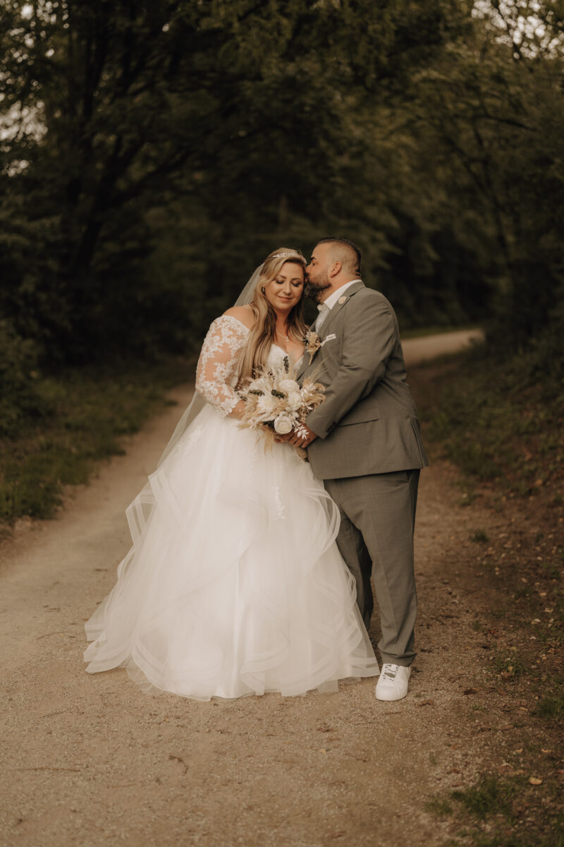 Brautpaar steht vor dem Wald, er küsst sie auf die Stirn, Hochformat warm