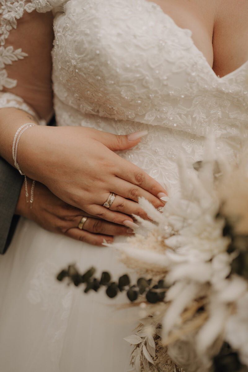 Foto Nahaufnahme Kleid, Hände vom Brautpaar auf dem Kleid übereinander gelegt und Brautstrauß auf rechts unten unscharf warm Hochformat