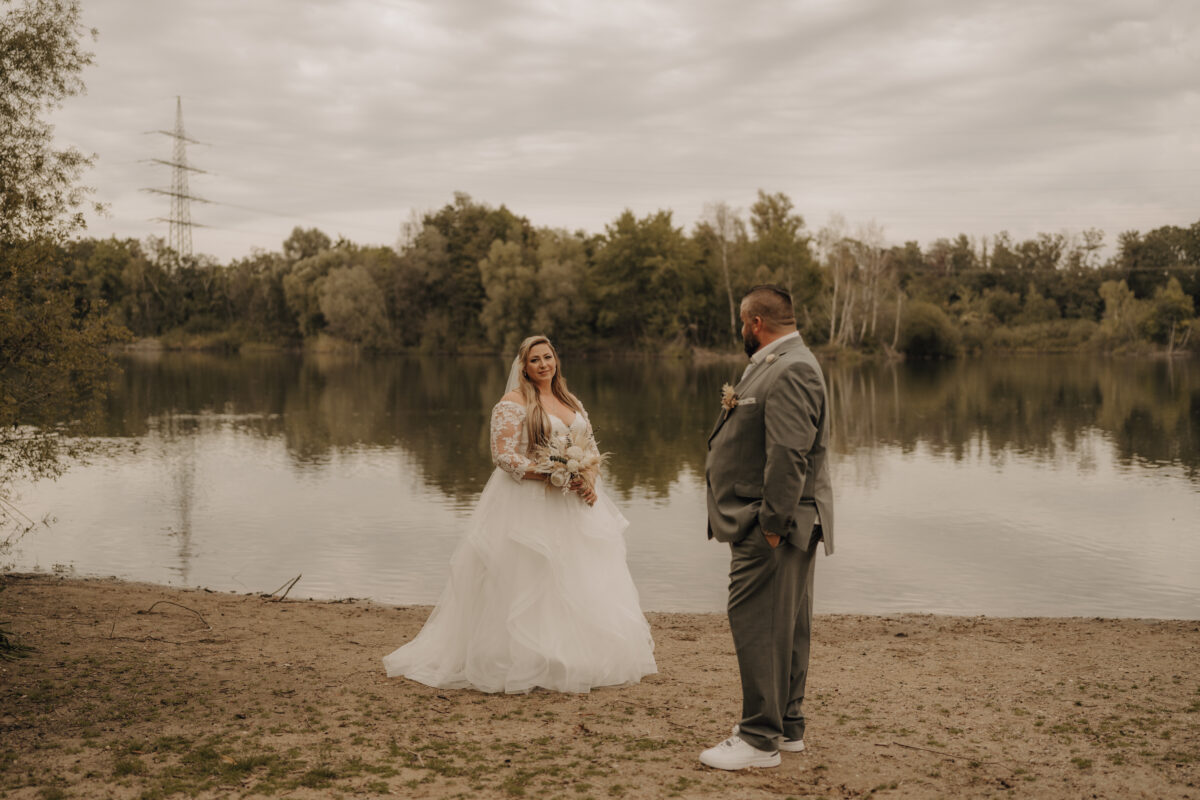 Foto Braut steh hinten und schaut nach vorne, Bräutigam steht vorne mit dem Rücken zu uns am Silbersee warm Querformat