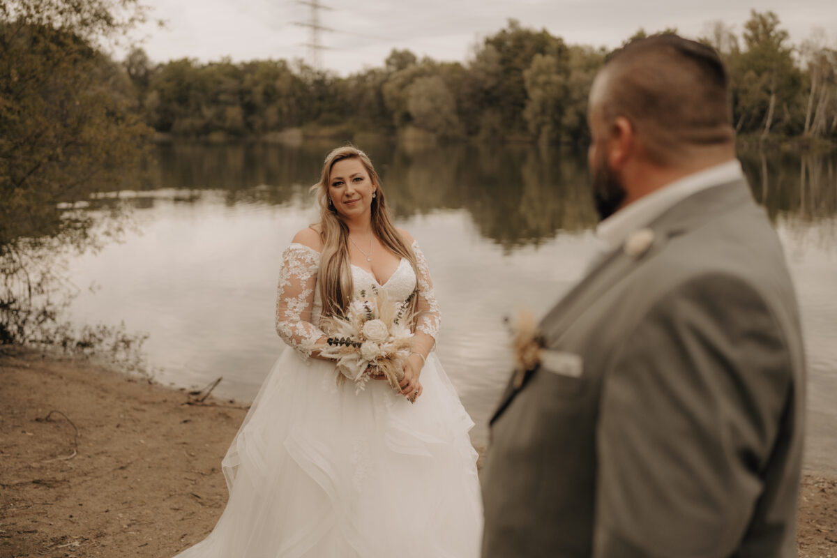 Bräutigam steh im Vordergrund mit Rücken zu uns verschwommen, Braut im Hintergrund schaut zu ihm nach vorne scharf, warm Querformat