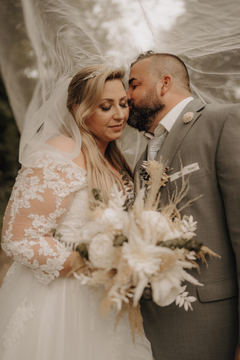 Bräutigam küsst Braut unterm Schleier auf die linke Wange, Querformat