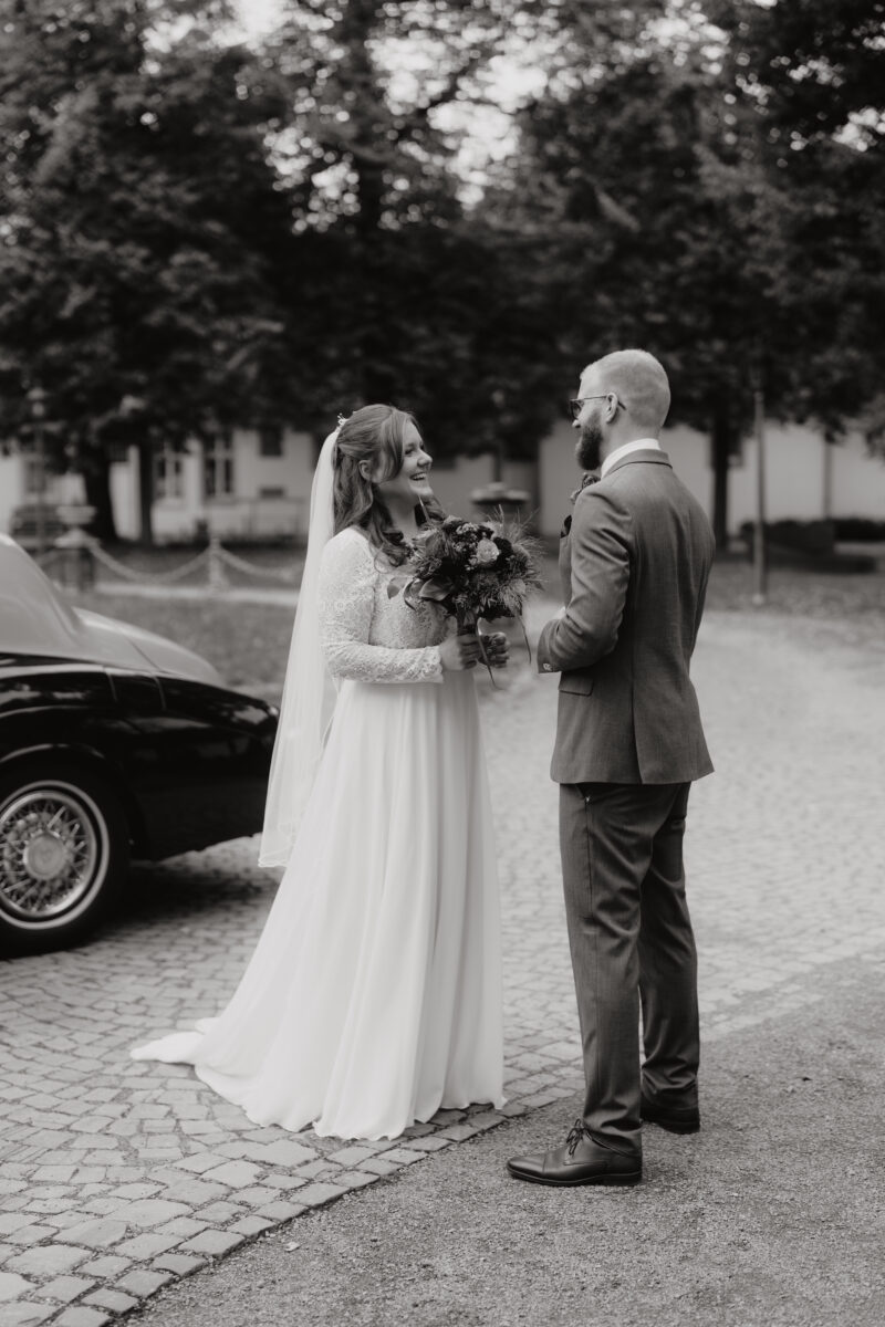 Hochzeitsportfolio, Die Braut und Bräutigam schauen sich lachend gegenseitig an im Hintergrund ein Auto, bild in Schwarz Weiß