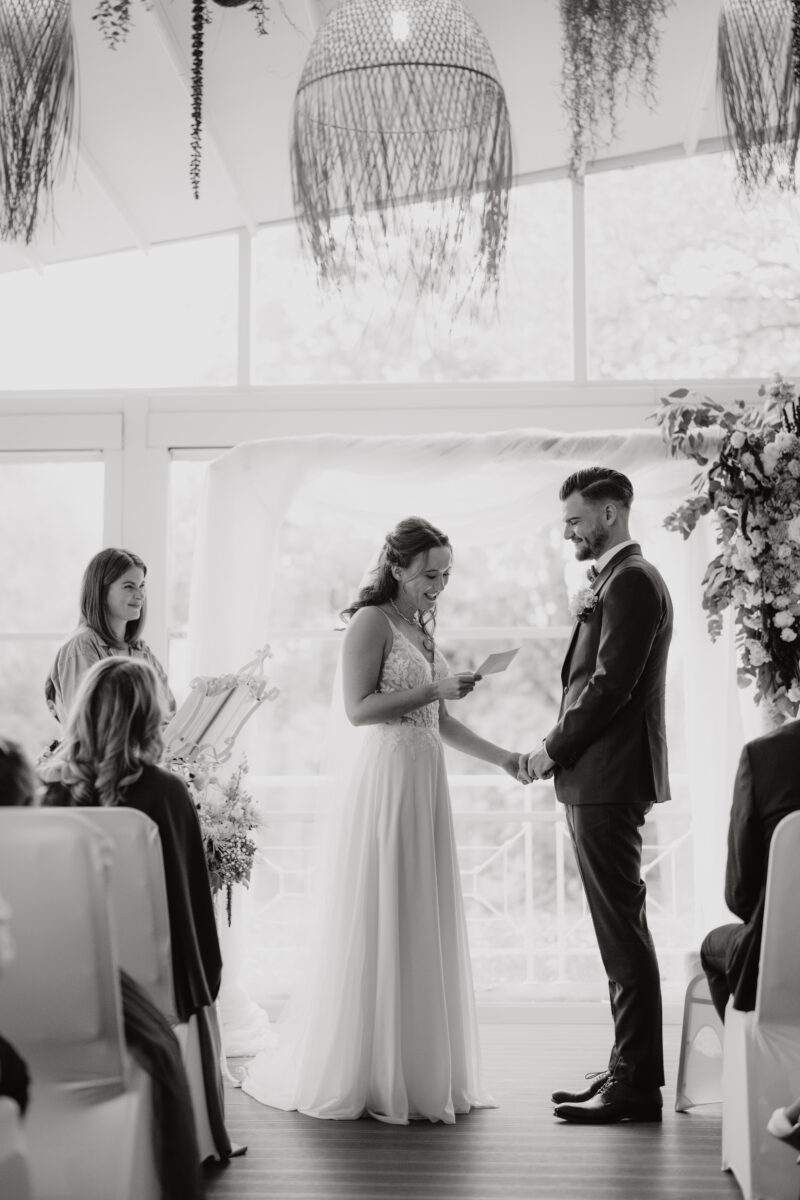Hochzeitsportfolio, Braut und Bräutigam stehen vorne während der Trauung, Braut hält die Hand des Bräutigams fest, während sie ihr Ehegelübde von einem Zettel vorliest, Bild in Schwarz Weiß