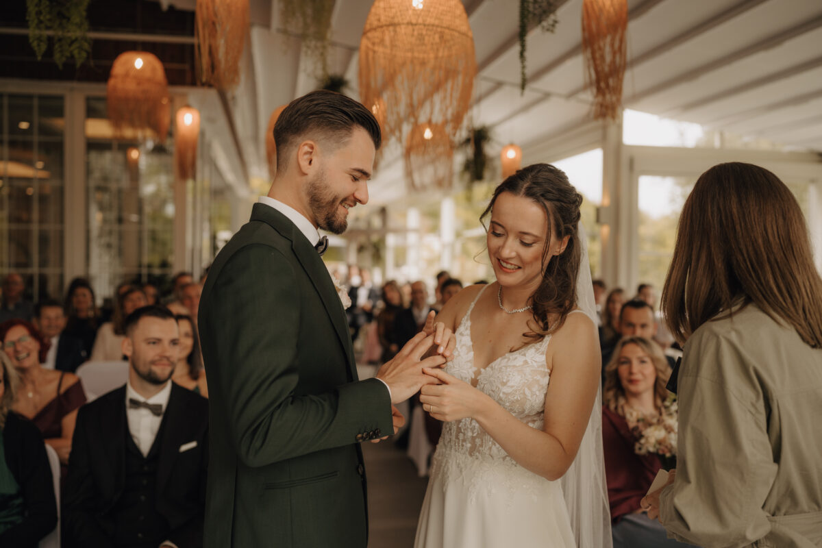 Hochzeitsportfolio, Braut und Bräutigam stehen vorne, vor den Gästen, Die Braut setzt den Ring auf den Finger des Bräutigams