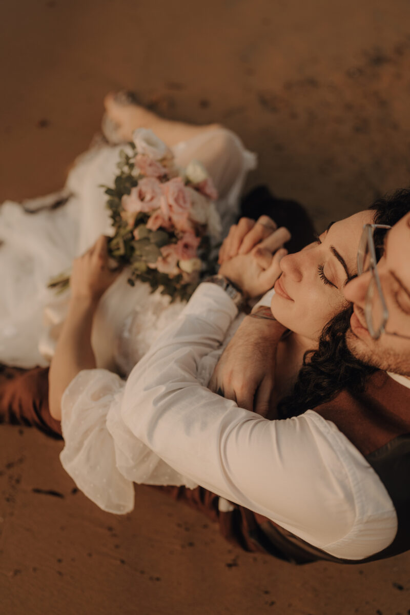 After Wedding Portfolio, Braut und Bräutigam liegen am Strand, die Braut in seinen Armen, und haben die Augen geschlossen
