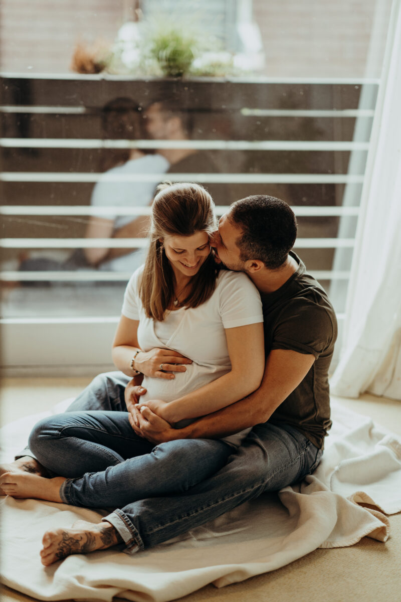 Babybauch Fotografie Portfolio werdendes Elternpaar posiert auf einer Decke vor einem Fenster