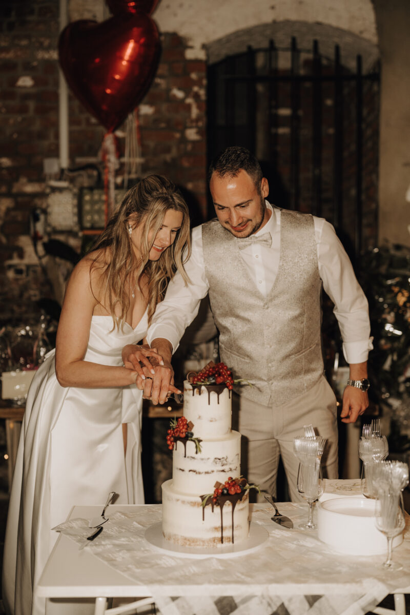 Hochzeitsportfolio, Braut und Bräutigam schneiden zusammen die Hochzeitstorte an, in der Hochzeitslocation