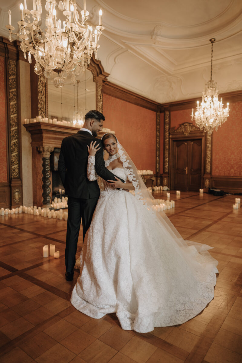 After Wedding Portfolio, Bräutigam steht rücklings zur Kamera, die Braut an seiner Seite steht in Richtung Kamera und hält die Schulter ihres Mannes, sie befinden sich in einem roten Ballsaal