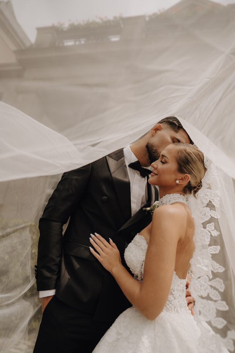 After Wedding Portfolio, Braut und Bräutigam stehen eng beieinander und der Bräutigam gibt der Braut einen Kuss auf die Stirn, über ihre Köpfe ist der Schleier der Braut gelegt