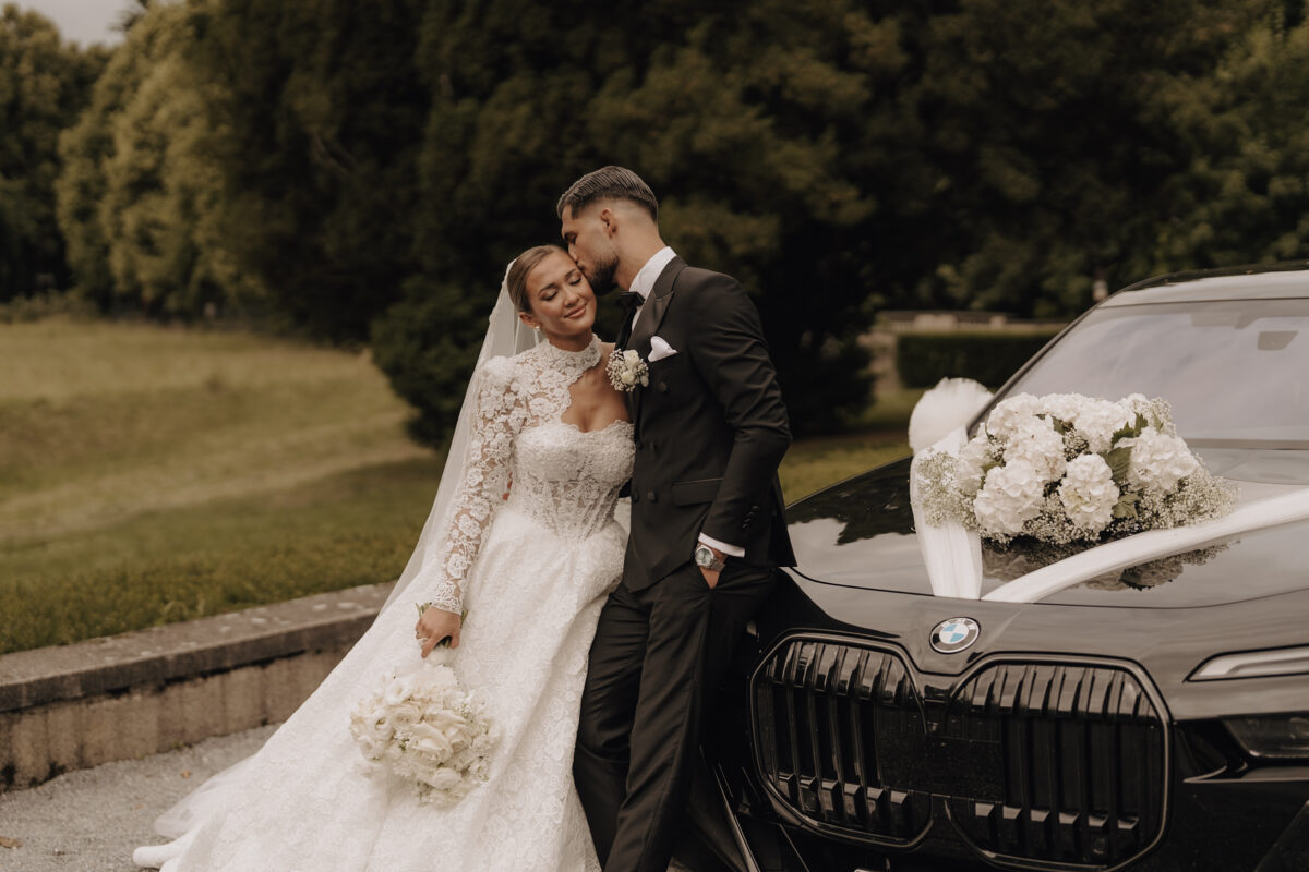 After Wedding Portfolio, Braut und Bräutigam lehnen an der Motorhaube eines schwarzen Autos, der Bräutigam gibt seiner Braut einen Kuss auf die Stirn