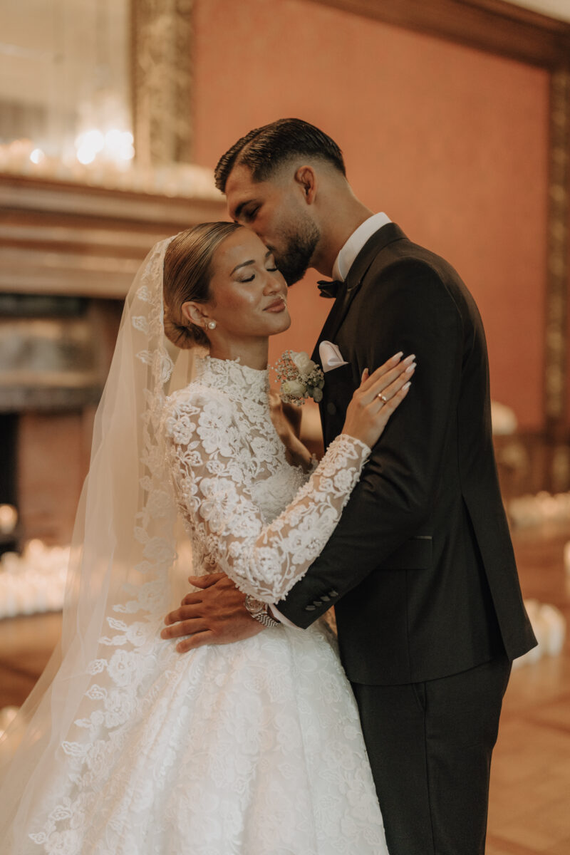 After Wedding Portfolio, Braut und Bräutigam stehen eng voreinander in einem roten Ballsaal, der Mann gibt seiner Frau eine Kuss auf die Stirn