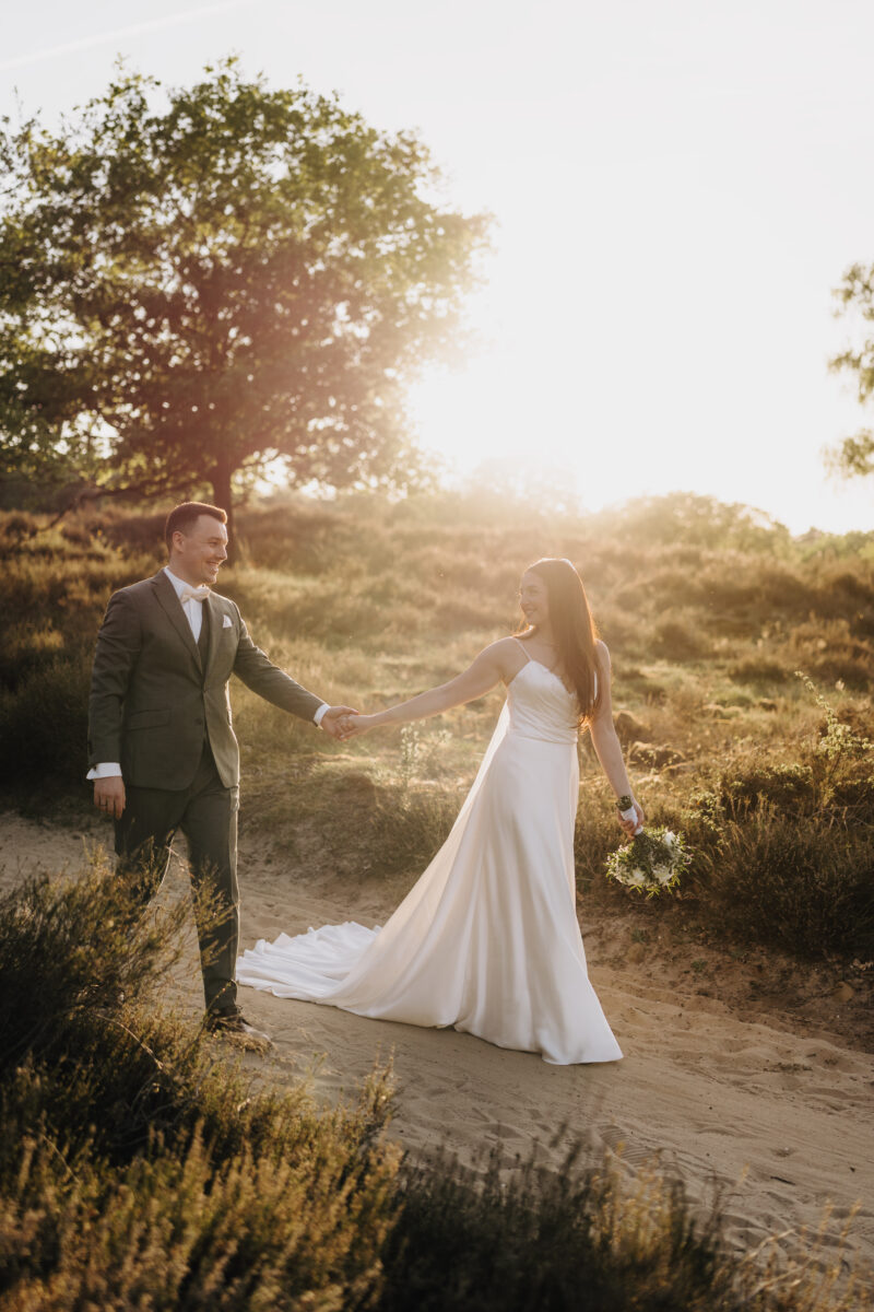 After Wedding Portfolio, Braut führt ihren Bräutigam, durch die Heide, im Hintergrund geht die Sonne unter