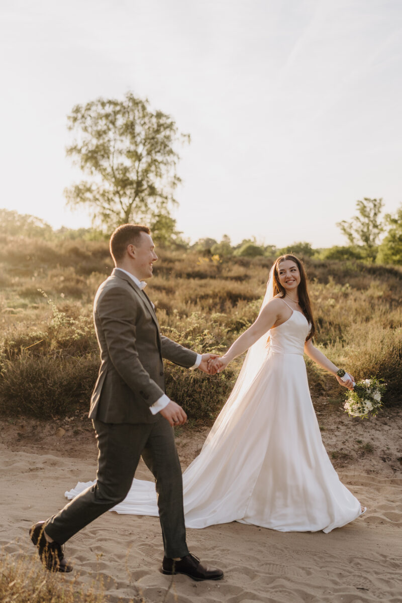After Wedding Portfolio, Braut und Bräutigam laufen händehaltend einen Weg in der Heide entlang und schauen sich glücklich an