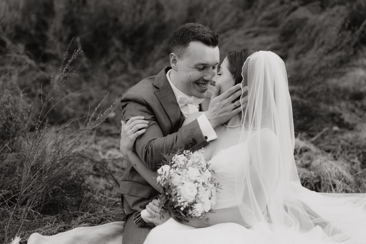 After Wedding Portfolio, Braut und Bräutigam sitzen in der Heide, der Bräutigam hält das Gesicht der Braut in den Händen und lacht sie an, Foto in schwarz-weiß