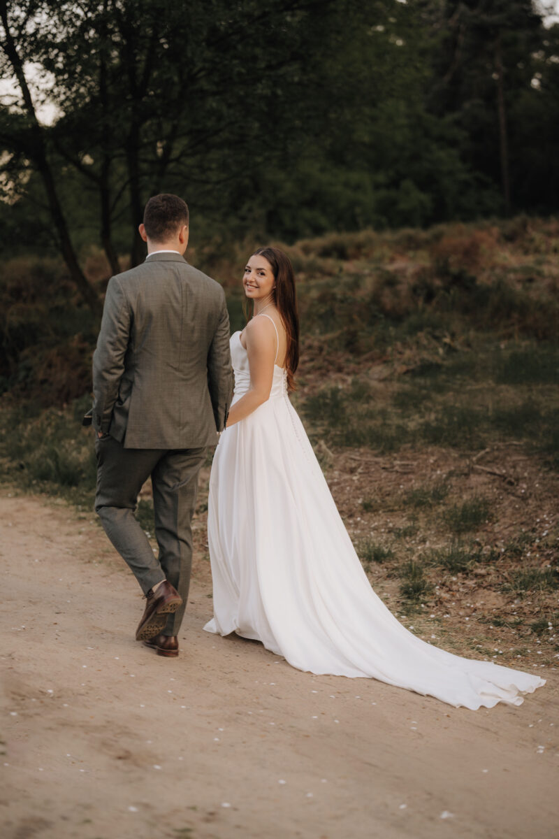 After Wedding Portfolio, Braut und Bräutigam laufen Hand in Hand einen Weg in der Natur entlang, die Braut schaut über die Schulter zurück in die Kamera