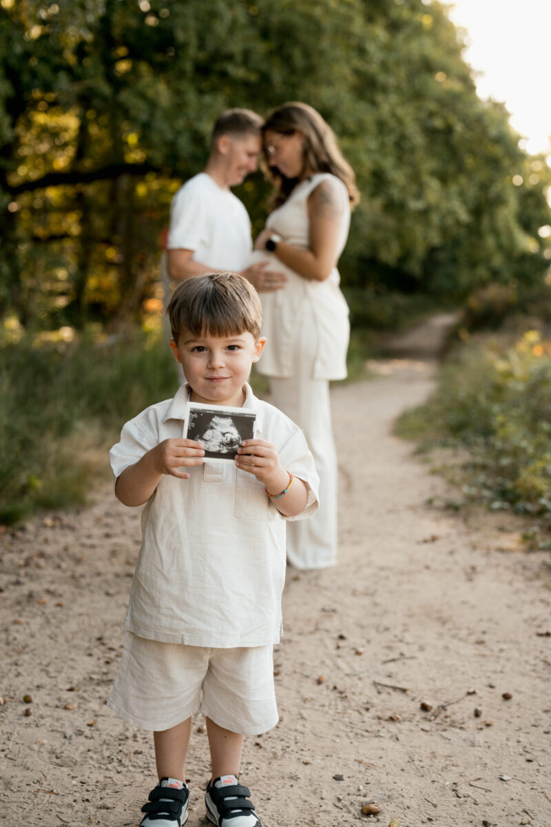 Babybauch Fotografie Portfolio Elternpaar posiert in der Natur Sohn läuft mit Ultraschallbild auf die Kamera zu
