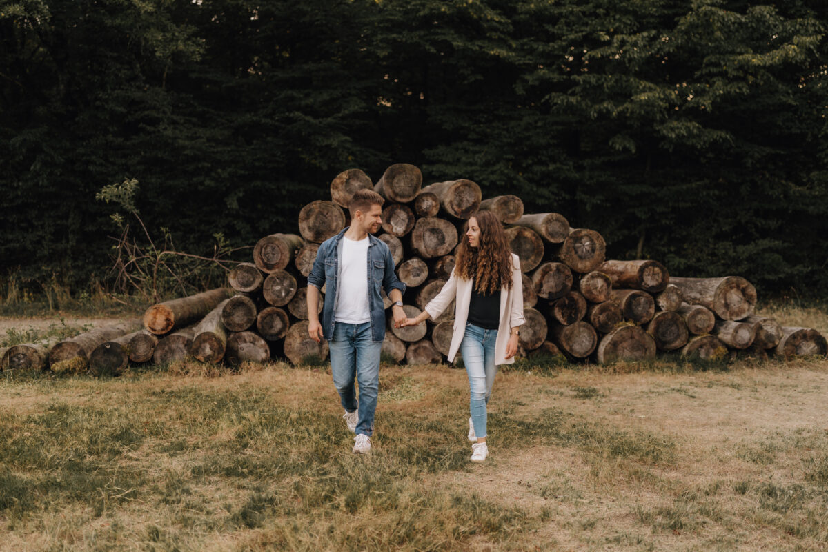 Paar schlendert Händchen haltend auf die Kamera zu, im Hintergrund sind gestapelte Baumstämme