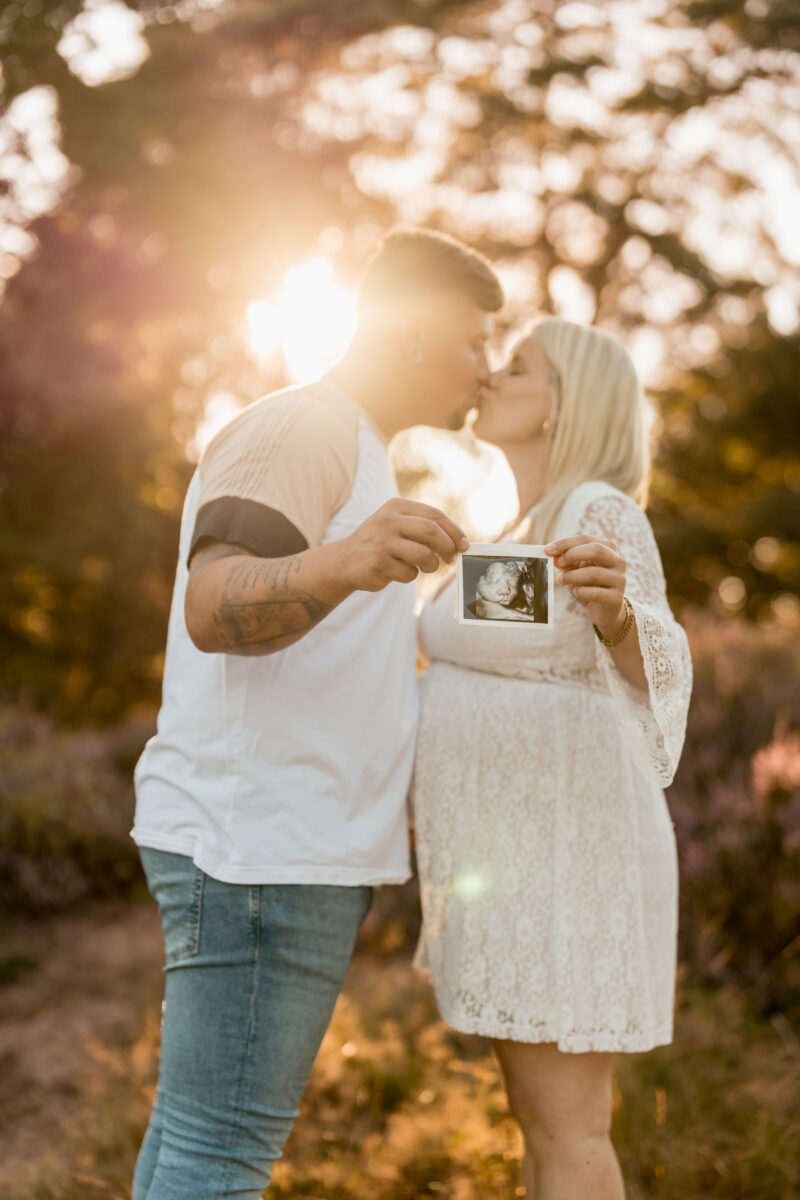 Babybauch Fotografie Portfolio junges glückliches Paar in der Natur zeigt Ultraschallbild in die Kamera und küsst sich